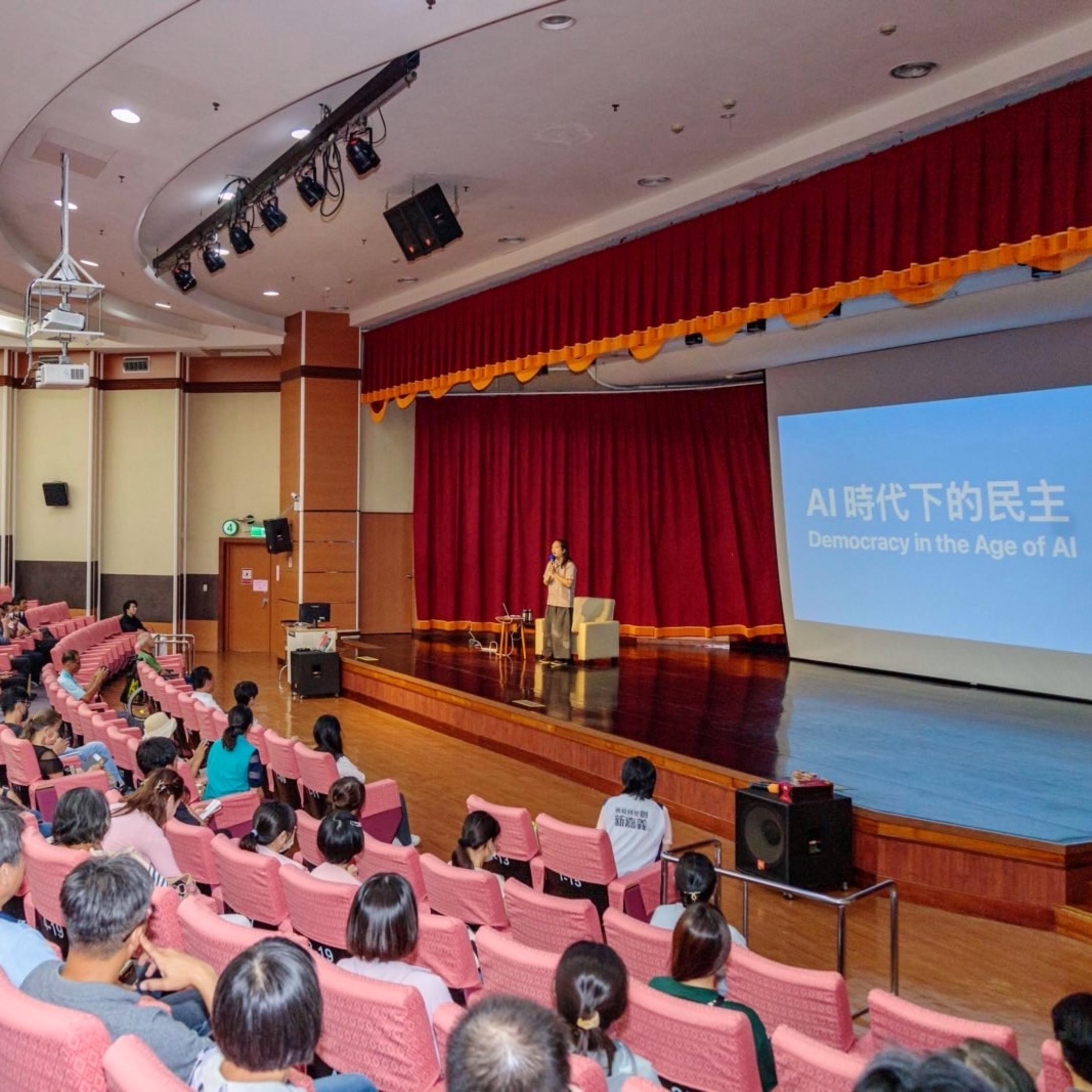 Audrey Tang on stage giving a speech in front of an audience, with iA Presenter in the background